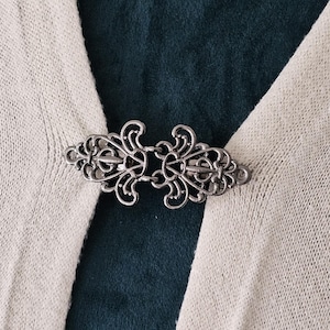 May include: A silver metal decorative clasp with a filigree design. The clasp is attached to a beige knitted fabric.