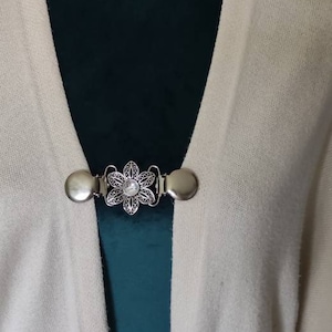 May include: A white cardigan with a silver filigree brooch featuring a clear rhinestone in the centre.