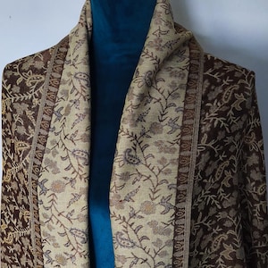 May include: A brown and beige shawl with floral and paisley patterns. The shawl is draped over a mannequin, showcasing the intricate designs and colour variations. The textile features detailed borders and a soft, flowing texture.