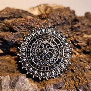 Könnte beinhalten: Ein silberfarbener Mandala-Ring mit filigranen Details. Der Ring ist aus Metall und hat ein kreisförmiges Design mit einem floralen Muster.