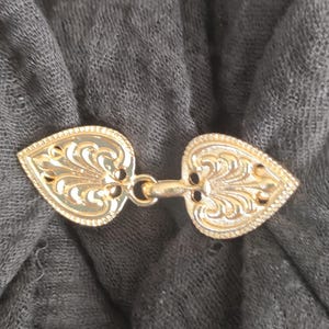 May include: A gold-coloured heart-shaped clasp with intricate details. The clasp is made of metal and has a decorative design.