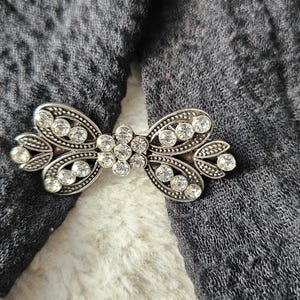 Peut inclure: Broche en métal argenté en forme de nœud, ornée de nombreux strass clairs et étincelants. La broche est posée sur un tissu gris foncé, créant un contraste qui met en valeur les détails complexes du bijou.