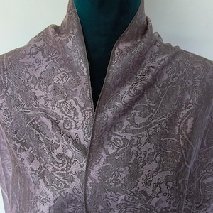 Könnte beinhalten: Ein mauvefarbener Schal mit Paisley-Muster, der über einer Schaufensterpuppe drapiert ist. Das Design des Schals zeigt ein detailliertes, sich wiederholendes Muster in Mauve- und Grautönen. Die Schaufensterpuppe hat einen dunkelgrünen Samtkragen.