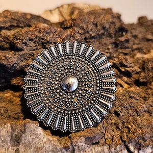 Könnte beinhalten: Ein silberfarbener, kreisförmiger Anhänger mit einem detaillierten, strukturierten Design. Der Anhänger hat einen zentralen, erhabenen Kreis, der von kleineren, perlenbesetzten Details und spitzen Kanten umgeben ist. Der Schmuck wird auf einem Stück strukturiertem, braunem Holz präsentiert.