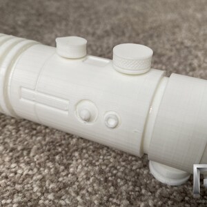 May include: White plastic lightsaber prop with a textured surface and two round buttons. The lightsaber is on a brown carpet.