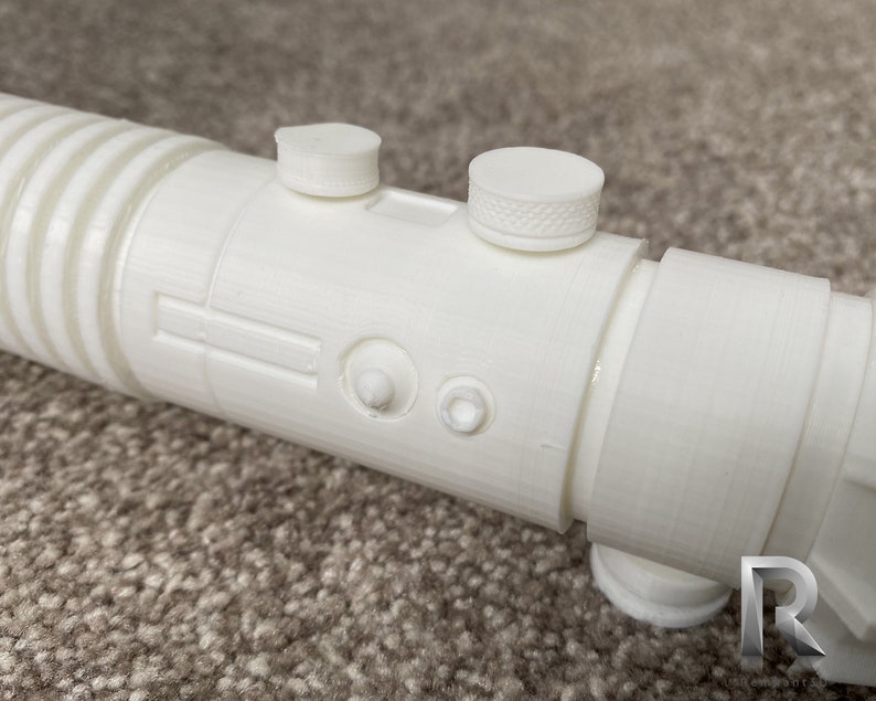 May include: White plastic lightsaber prop with a textured surface and two round buttons. The lightsaber is on a brown carpet.