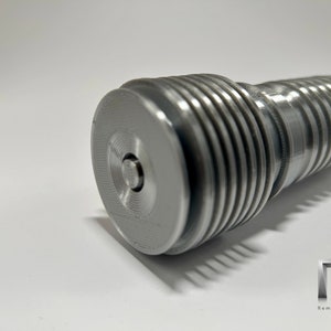 May include: Close-up of a silver metal lightsaber hilt with a textured, ribbed design. The hilt has a small, circular button on the end.