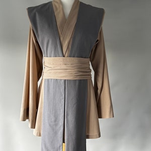 May include: A tan and gray costume displayed on a mannequin. The costume features a long-sleeved tan tunic, a gray overcoat, and a tan wrap belt. The overcoat has a V-neck design. The costume is designed to be worn by a person.
