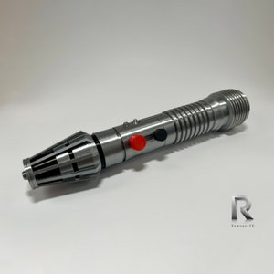 May include: A silver lightsaber prop with a black and red button. The lightsaber has a textured, cylindrical handle and a pointed tip.