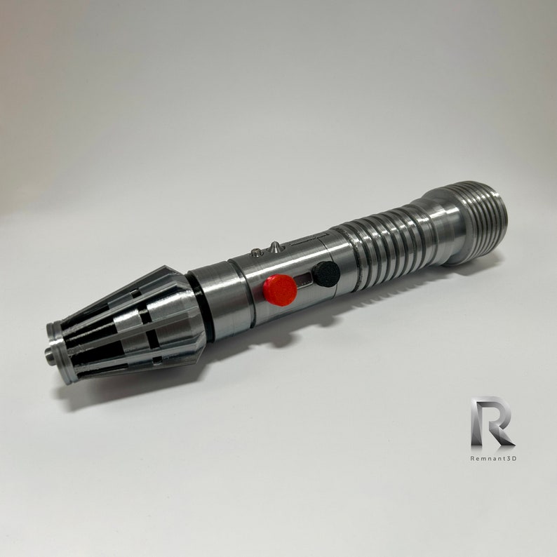 May include: A silver lightsaber prop with a black and red button. The lightsaber has a textured, cylindrical handle and a pointed tip.
