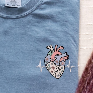 May include: Blue jean colored t-shirt with a floral anatomical heart design and a heartbeat line. The shirt has a crew neck and short sleeves. The text "Super Soft" is in the upper right corner.