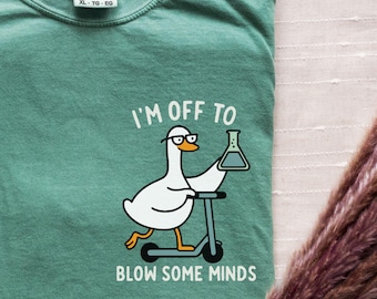 May include: A light green t-shirt featuring a graphic of a white goose wearing glasses, riding a scooter, and holding a laboratory flask. The text above and below the goose reads, I'M OFF TO BLOW SOME MINDS. The shirt has a crew neckline.