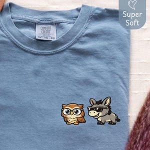 May include: A blue jean colored t-shirt with a small embroidered design of an owl wearing glasses and a donkey wearing sunglasses. The shirt has a tag that says "Comfort Colors". The text "Blue Jean" is in the lower left corner.
