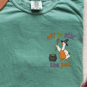 May include: A light green t-shirt featuring a graphic of a white goose wearing a black witch hat and an orange apron. The goose holds a wooden spoon over a black cauldron with green liquid. The text reads off to stir the pot in colorful letters.