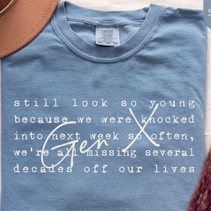 Flat lay of a Comfort Colors “Blue Jean” super soft tee on a light fabric background with a tan hat and pampas grass. White text reads: “still look so young because we were knocked into next week so often, we’re all missing several decades off our lives.” Funny Gen X nostalgia humor shirt for everyday wear, birthdays, and reunions - designed by PhraseArtStudio, Still Kickin Club.