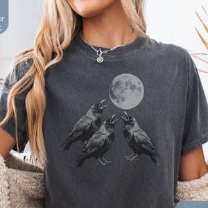 May include: A charcoal gray t-shirt featuring a graphic print of three black crows looking up toward a full moon. The shirt has a crew neckline and short sleeves. A small label in the top left corner reads Super Soft, and a label in the bottom right reads Pepper.