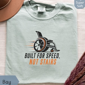 May include: A light green t-shirt with a black and orange graphic of a wheelchair and the text "BUILT FOR SPEED, NOT STAIRS". The shirt is a soft material and has a round neckline.