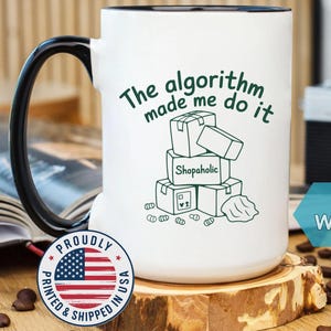 May include: White 15 oz coffee mug with a black handle and rim. The mug features the text "The algorithm made me do it" and a graphic of stacked boxes labeled "Shopaholic". A sticker with the American flag is visible.