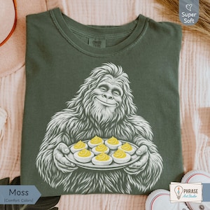 May include: A moss green crew neck t-shirt featuring a white graphic illustration of a smiling Bigfoot holding a platter of six deviled eggs with yellow yolks. The shirt has a tag on the inside collar that reads Comfort Colors.