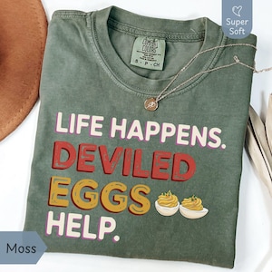 May include: A moss green t-shirt with the text "LIFE HAPPENS. DEVILED EGGS HELP." in white, red, and yellow. The shirt has a crew neck and short sleeves. A gold necklace with a pendant is draped on the shirt.