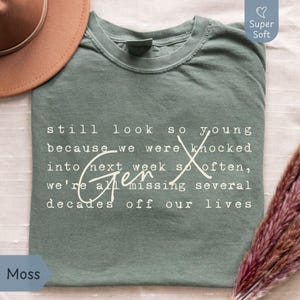 Flat lay of a Comfort Colors “Moss” green super soft tee on a light fabric background with a tan hat and pampas grass. White Gen X nostalgia quote in typewriter font with script “Gen X” reads: “still look so young because we were knocked into next week so often, we’re all missing several decades off our lives.” Funny Gen X humor shirt for everyday wear, birthdays, and reunions - designed by PhraseArtStudio, Still Kickin Club.