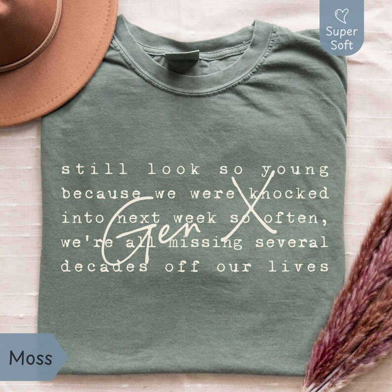 Flat lay of a Comfort Colors “Moss” green super soft tee on a light fabric background with a tan hat and pampas grass. White Gen X nostalgia quote in typewriter font with script “Gen X” reads: “still look so young because we were knocked into next week so often, we’re all missing several decades off our lives.” Funny Gen X humor shirt for everyday wear, birthdays, and reunions - designed by PhraseArtStudio, Still Kickin Club.