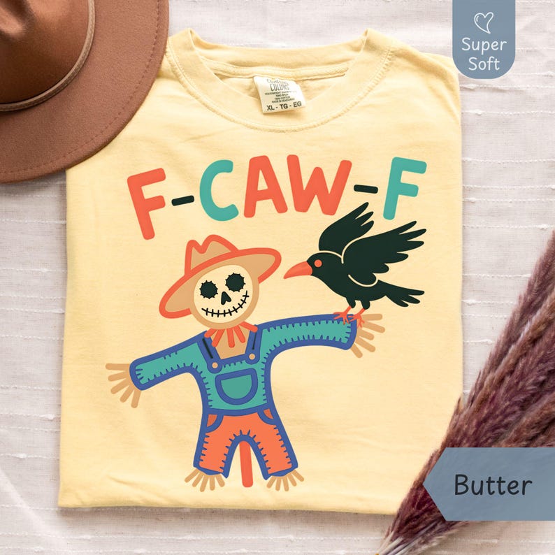 Butter yellow Comfort Colors T-shirt featuring a playful “F-CAW-F” design with a cartoon scarecrow and a black crow perched on its arm. Laid flat on a neutral background with a brown hat for a cozy, rustic vibe. Made from super soft cotton, this graphic tee is ideal for fall wear, Halloween events, or gifting to anyone who loves lighthearted humor and seasonal designs. Available in multiple sizes through Phrase Art Studio’s Etsy shop.