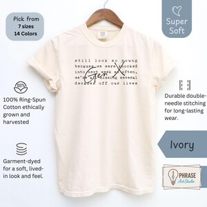 Product photo of an Ivory Comfort Colors garment-dyed tee on a wooden hanger against a white backdrop. Gen X humor text design reads “still look so young…” in typewriter style with script Gen X. Callouts show super soft feel, 100% ring-spun cotton, double-needle stitching, and choice of 7 sizes and 14 colors. Funny 90s nostalgia shirt for everyday wear, birthdays, and reunions - designed by PhraseArtStudio, Still Kickin Club.