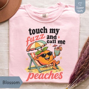 Blossom pastel pink-colored Comfort Colors shirt featuring a colorful retro cartoon peach sitting under a beach umbrella reading 'touch my fuzz and call me peaches' for a sassy summer vibe. Designed by experts at PhraseArtStudio - to solve the problem of finding a funny, zero-filter women's joke shirt.