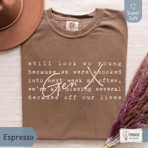 Flat lay of a super soft Comfort Colors “Espresso” brown tee on a light fabric background with a tan hat and pampas grass, plus a small PhraseArtStudio logo. Cream Gen X nostalgia quote in typewriter font with script “Gen X” reads: “still look so young because we were knocked into next week so often, we’re all missing several decades off our lives.” Funny Gen X humor shirt for everyday wear, birthdays, and reunions - designed by PhraseArtStudio, Still Kickin Club.