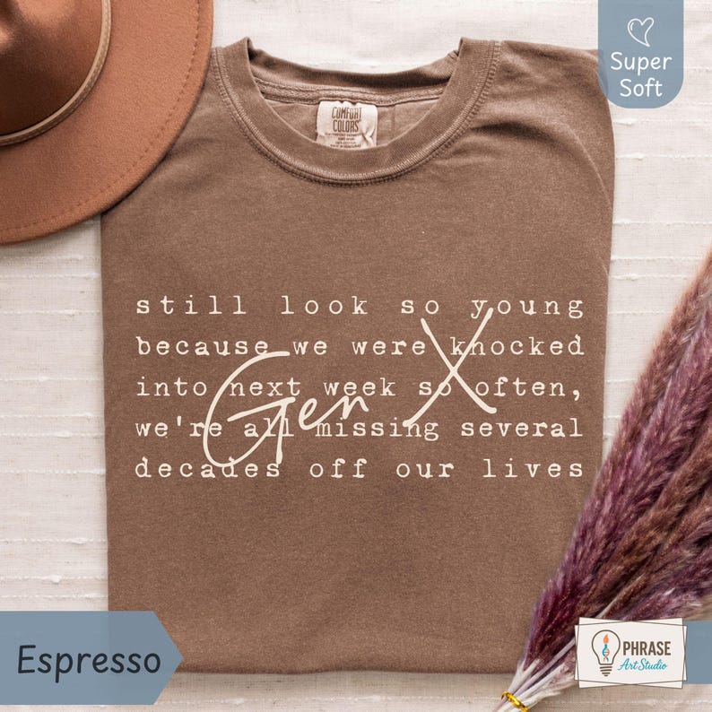 Flat lay of a super soft Comfort Colors “Espresso” brown tee on a light fabric background with a tan hat and pampas grass, plus a small PhraseArtStudio logo. Cream Gen X nostalgia quote in typewriter font with script “Gen X” reads: “still look so young because we were knocked into next week so often, we’re all missing several decades off our lives.” Funny Gen X humor shirt for everyday wear, birthdays, and reunions - designed by PhraseArtStudio, Still Kickin Club.