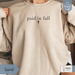 Könnte beinhalten: Ein sandfarbenes Sweatshirt mit Rundhalsausschnitt und dem schwarzen Schriftzug "paid in full" mittig auf der Brust, wobei sich direkt unter dem Text ein kleines schwarzes Kreuzsymbol befindet. Das Kleidungsstück hat lange Ärmel und Rippbündchen.