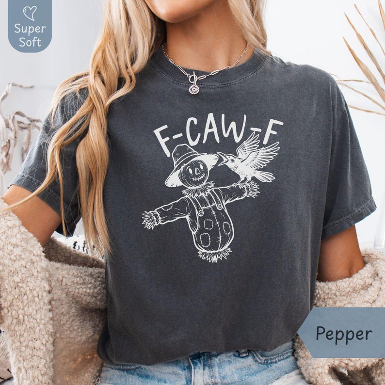 Pepper Comfort Colors T-shirt for women featuring a white “F-CAW-F” graphic of a scarecrow with a crow perched on its arm. Modeled indoors with a cozy sherpa cardigan, this cropped, relaxed-fit tee has a casual fall vibe. Made from super soft cotton, it’s perfect for autumn outfits, Halloween season, or gifting to someone who loves playful, rustic designs with a touch of sass. Available in multiple sizes through Phrase Art Studio’s Etsy shop.