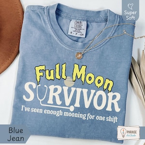 Könnte beinhalten: Ein jeansblaues T-Shirt mit dem Text Full Moon SURVIVOR und I've seen enough mooning for one shift in gelber und weißer Schrift. Das Design enthält eine Grafik eines Stethoskops und eines Mondes. Das T-Shirt hat einen Rundhalsausschnitt.