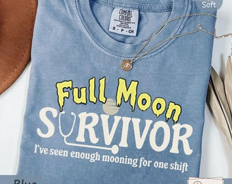 Può includere: Una t-shirt color blu jeans con il testo Full Moon SURVIVOR e I've seen enough mooning for one shift in lettere gialle e bianche. Il design include un grafico di uno stetoscopio e una luna. La t-shirt ha uno scollo rotondo.