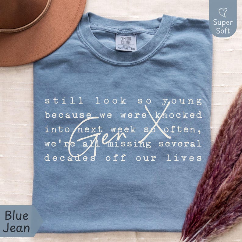 Flat lay of a Comfort Colors “Blue Jean” super soft tee on a light fabric background with a tan hat and pampas grass. White text reads: “still look so young because we were knocked into next week so often, we’re all missing several decades off our lives.” Funny Gen X nostalgia humor shirt for everyday wear, birthdays, and reunions - designed by PhraseArtStudio, Still Kickin Club.