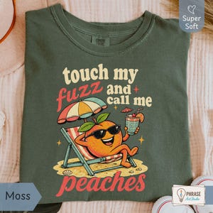 Moss sage green-colored Comfort Colors shirt featuring a vibrant 80s style cartoon peach with sunglasses reading 'touch my fuzz and call me peaches' for an ironic retro meme vibe. Designed by experts at PhraseArtStudio - to solve the problem of finding unique, sarcastic adult humor gifts.
