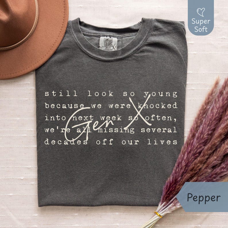 Flat lay of a super soft Comfort Colors “Pepper” dark gray tee on a light fabric background with a tan hat and pampas grass. Cream Gen X nostalgia quote in typewriter font with script “Gen X” reads: “still look so young because we were knocked into next week so often, we’re all missing several decades off our lives.” Funny Gen X humor shirt for everyday wear, birthdays, and reunions - designed by PhraseArtStudio, Still Kickin Club.