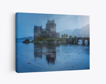 May include: A gray stone castle on a small island with a stone bridge leading to it. The castle is reflected in the still water of the loch. The sky is a light blue with some clouds.