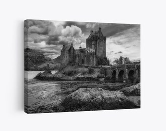May include: Black and white canvas print of a medieval castle on a rocky island. The castle has multiple towers and is connected to the mainland by a stone bridge. The sky is filled with dramatic clouds.