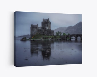 May include: A gray stone castle with a bridge in front of it. The castle is on a small island in a body of water. The water is calm and reflects the sky and the castle. The sky is overcast and gray.