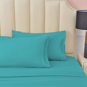 May include: A teal blue bed sheet set with two matching pillowcases. The bed sheet set is on a white headboard with a decorative design.