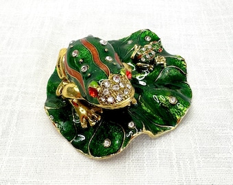 May include: A green and gold frog trinket box with a jeweled design. The frog is sitting on a green leaf with a smaller frog on the leaf.
