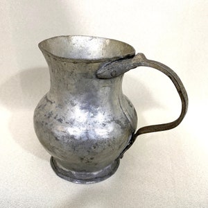 Peut inclure: Une cruche en argent avec un corps arrondi et une anse incurvée. La cruche a un aspect usé et vieilli.
