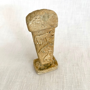 May include: A small, beige stone sculpture of a column with carved animal figures. The column has a square base and a rounded top. The animals are carved in relief and appear to be a cow and a deer.