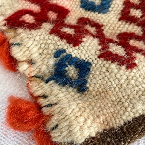 Pu&ograve; includere: Primo piano di un arazzo tessuto con filo rosso, blu e bianco. L'arazzo ha un bordo a frangia e una superficie testurizzata.