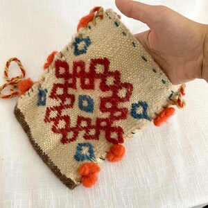 Pu&ograve; includere: Una piccola borsa intrecciata di colore crema con un motivo geometrico in rosso, blu e arancione. La borsa ha una chiusura a coulisse ed &egrave; decorata con pompon.