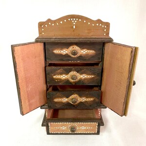 May include: A wooden jewelry box with four drawers and a hinged door. The drawers and door are decorated with intricate patterns of inlaid bone or shell. The box is painted brown and has a natural wood finish.