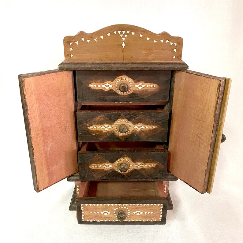 May include: A wooden jewelry box with four drawers and a hinged door. The drawers and door are decorated with intricate patterns of inlaid bone or shell. The box is painted brown and has a natural wood finish.