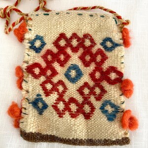 Pu&ograve; includere: Una piccola borsa intrecciata di colore crema con un motivo geometrico in rosso, blu e marrone. La borsa ha una chiusura a coulisse ed &egrave; decorata con pompon rossi.